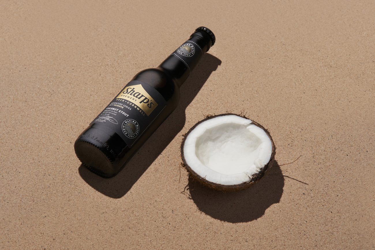 Lifestyle photo of a bottle of Sharp's Brewery Coconut Stout on sand next to half a coconut