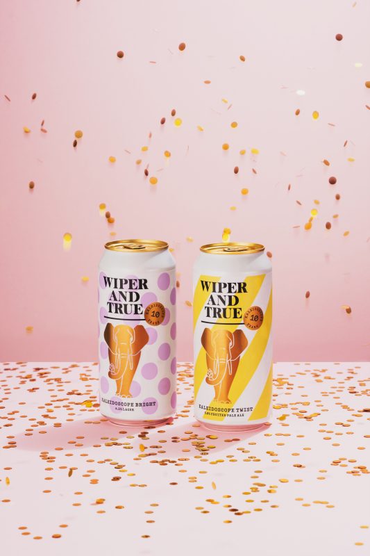 A studio photograph of Wiper and True Kaleidoscope 10th anniversary beers - a fruited pale ale and a lager - on a pink background with gold confetti